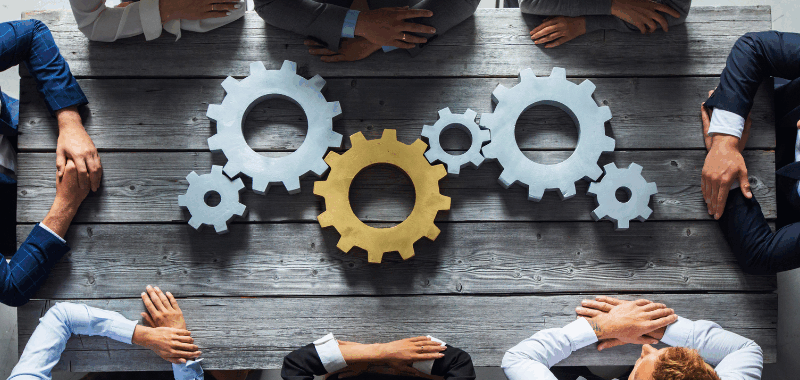 Equipo de trabajo reunido alrededor de engranajes sobre una mesa, representando la integración entre gestión de proyectos y análisis de negocio para optimizar la operación.