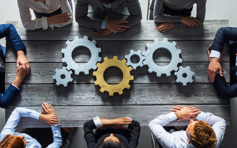 Equipo de trabajo reunido alrededor de engranajes sobre una mesa, representando la integración entre gestión de proyectos y análisis de negocio para optimizar la operación.