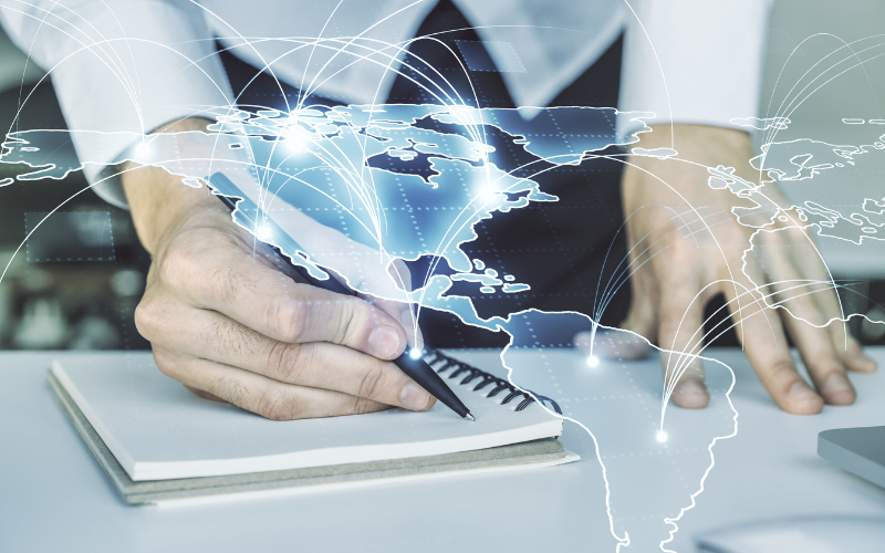 Manos escribiendo en un cuaderno mientras un mapa digital global muestra conexiones entre países, representando la armonización normativa internacional y la transformación regulatoria y tecnológica del sector financiero en México